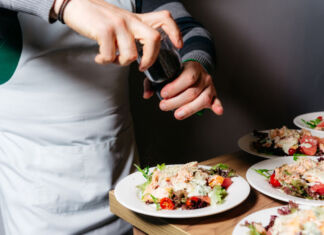 Ein Koch würzt mit einer Gewürzmühle den auf den weißen Tellern angerichteten Salat. Er trägt eine graue Schürze und arbeitet an einem Holztisch.
