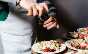 Ein Koch würzt mit einer Gewürzmühle den auf den weißen Tellern angerichteten Salat. Er trägt eine graue Schürze und arbeitet an einem Holztisch.
