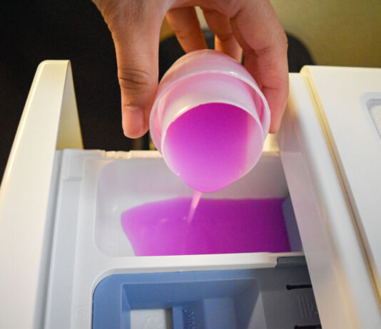 Eine Frau kippt flüssiges Waschmittel oder Weichspüler in Pink in die Einspülkammer der Waschmaschine. Die Hand ist im Vordergrund, ebenso die Schublade.
