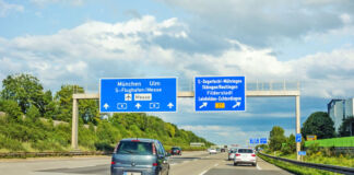 Eine sonnige Autobahn in Deutschland im Sommer, auf der zahlreiche Autos und Kleinwagen fahren. Über ihnen hängen einige verkehrsweisende Streckenschilder.