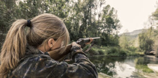 Eine Frau mit einer braun-grünen Jacke und einem Zopf ist von hinten zu sehen, während sie mit einem Gewehr im Wald steht. Die Waffe ist auf ein Tier gerichtet, welches sie jagt.