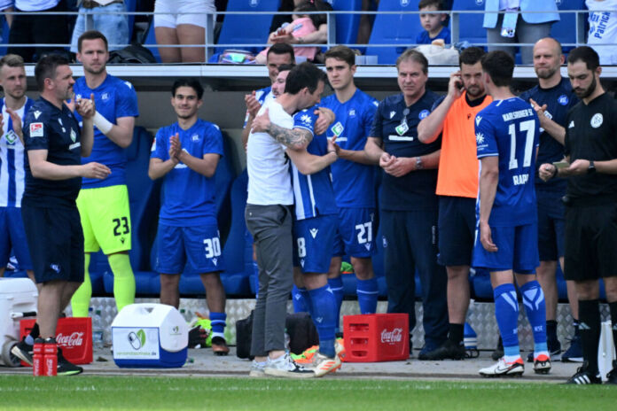 Die Spieler des Karlsruher SC umarmen ihren Trainer Christian Eichner und applaudieren ihm nach einem Spiel und dem Ende der Saison. Sie stehen zusammen in einem Stadion direkt am Spielfeld. Jetzt kommt ein Bayern-Star zum KSC.