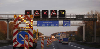 Reger Verkehr auf der Autobahn. Autos fahren unter einer Signalbrücke mit verschiedenen blinkenden Verkehrszeichen drunter. Darunter befindet sich ein mysteriöser Pfeil, der auf eine Ableitung oder ähnliches hinzuweisen scheint.