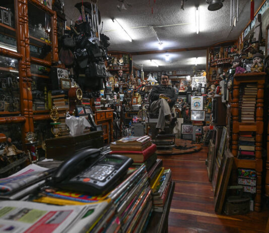 Ein Barbier sitzt in seinem Barber-Shop. Im Vordergrund liegen Magazine und ein Telefon. Der Laden ist voll mit alten Schränken, Büchern, Puppen, Kameras. Der Friseur-Salon erinnert an ein Antiquitäten-Geschäft.