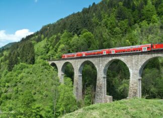 Eine rote Bahn fährt an einem schönen Tag mit blauem Himmel über eine hohe Brücke mitten durch den Wald in Baden-Württemberg