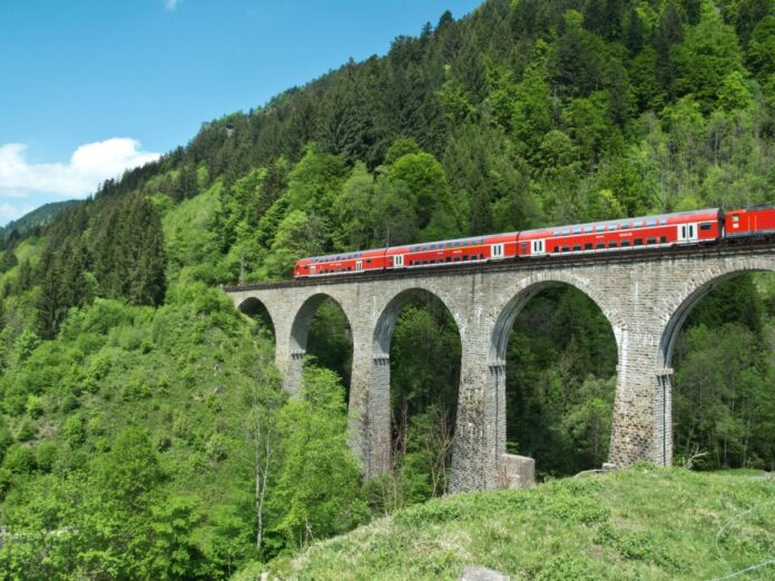 Eine rote Bahn fährt an einem schönen Tag mit blauem Himmel über eine hohe Brücke mitten durch den Wald in Baden-Württemberg