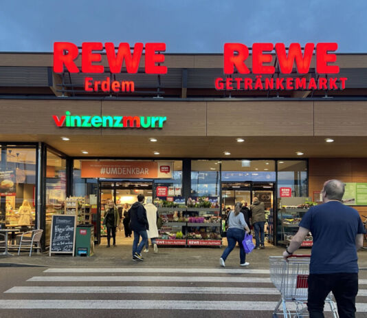 Ein beleuchteter riesiger Eingang eines Rewe Supermarktes mit einem Getränkemarkt von Rewe daneben. Vor der Filiale gehen einige Kunden über den Zebrastreifen und betreten den Supermarkt. Links befindet sich eine Bäckerei.