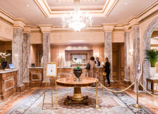 In der Lobby des Hotel Regent stehen zwei Gäste an der Rezeption, während der Empfangschef etwas sagt. Die Lobby ist sehr edel und ist in Gold und Marmor ausgestattet. In der Mitte befindet sich ein Tisch, der auf einem edlen Teppich steht.