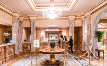 In der Lobby des Hotel Regent stehen zwei Gäste an der Rezeption, während der Empfangschef etwas sagt. Die Lobby ist sehr edel und ist in Gold und Marmor ausgestattet. In der Mitte befindet sich ein Tisch, der auf einem edlen Teppich steht.