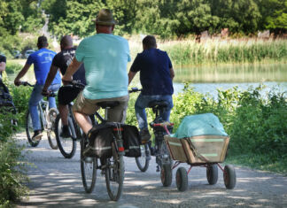 Mehrere Männer von hinten zu sehen fahren an einem sonnigen Tag Fahrrad entlang eines Sees. Der Fahrradfahrer rechts zieht einen Bollerwagen.