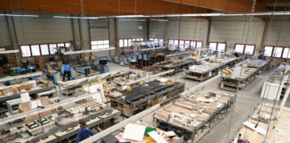 In einer riesigen Fabrikhalle stehen viele Arbeitstische nebeneinander, auf denen sich allerhand Werkzeug zum Herstellen befindet. Einige Mitarbeiter stehen mit Ohrschützern an den Tischen. Offensichtlich wird hier etwas aus Holz hergestellt.
