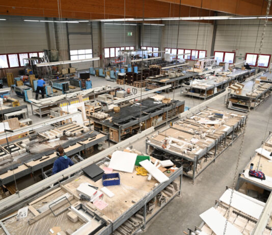 In einer riesigen Fabrikhalle stehen viele Arbeitstische nebeneinander, auf denen sich allerhand Werkzeug zum Herstellen befindet. Einige Mitarbeiter stehen mit Ohrschützern an den Tischen. Offensichtlich wird hier etwas aus Holz hergestellt.