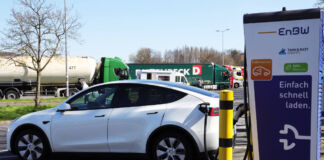 Ein E-Auto wird auf einem Parkplatz an einer E-Ladestation mit einem Kabel aufgeladen. Im Hintergrund befinden sich einige LKWs, die ebenfalls auf dem Parkplatz stehen.