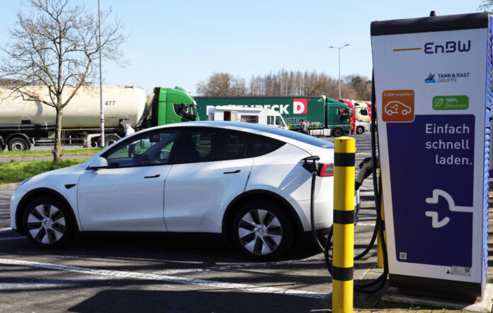 Ein E-Auto wird auf einem Parkplatz an einer E-Ladestation mit einem Kabel aufgeladen. Im Hintergrund befinden sich einige LKWs, die ebenfalls auf dem Parkplatz stehen.