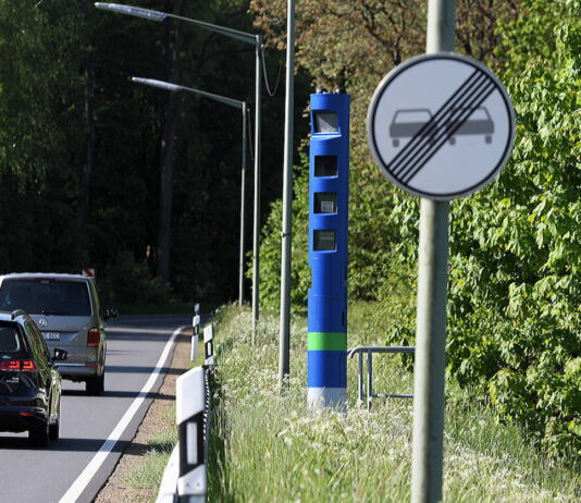 Am Straßenrand einer Bundesstraße oder Landstraße steht eine blaue Säule, eine Maut-Kontrollsäule. Im Vordergrund steht das Verkehrsschild "Überholverbot aufgehoben". Zwei Autos fahren in den mit Büschen und Bäumen bewachsenen Wald.
