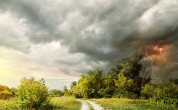 Ein mächtiges Gewitter zieht auf einer sommerlichen Landschaft mit Feldweg auf. Die Wolken sind düster und ein enormer Blitz entlädt sich in ihnen.