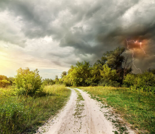 Ein mächtiges Gewitter zieht auf einer sommerlichen Landschaft mit Feldweg auf. Die Wolken sind düster und ein enormer Blitz entlädt sich in ihnen.