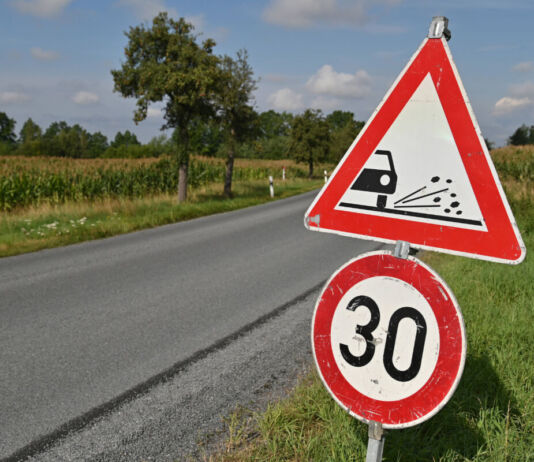 Auf einer Landstraße steht ein Verkehrszeichen, das die Geschwindigkeit auf 30 km/h begrenzt. Ursache ist der auf der Fahrbahn verteilte Rollsplitt. Am Straßenrand befinden sich Felder und Bäume.