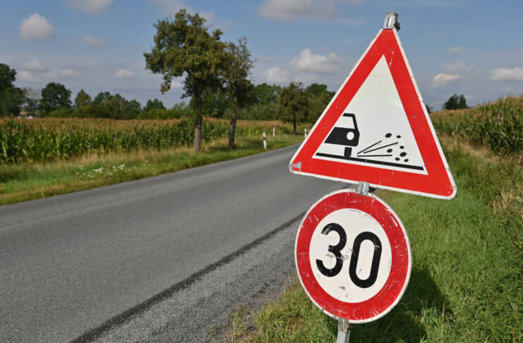 Auf einer Landstraße steht ein Verkehrszeichen, das die Geschwindigkeit auf 30 km/h begrenzt. Ursache ist der auf der Fahrbahn verteilte Rollsplitt. Am Straßenrand befinden sich Felder und Bäume.