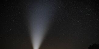 Ein Mann hält eine Taschenlampe auf freiem Feld in den Himmel der Nacht. Vereinzelte Sternenschnuppen machen sich am Himmelszelt breit.