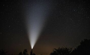 Ein Mann hält eine Taschenlampe auf freiem Feld in den Himmel der Nacht. Vereinzelte Sternenschnuppen machen sich am Himmelszelt breit.