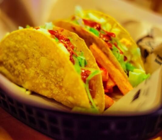 Kross gebackene und lecker gefüllte Tacos stehen auf einem Servierkorb. Unter ihnen liegt eine Serviette. Die Farbtöne sind in gelben Nuancen gehalten.