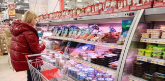 Eine Frau steht vor einem voll gepackten Kühlregal im Supermarkt. Sie trägt eine rote Jacke und wählt aus der Kühltheke Produkte aus. In ihrem Einkaufswagen befinden s ich bereits einige Lebensmittel.