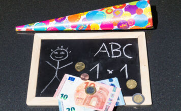 Eine kleine Tafel mit einem Strichmännchen und Schulaufgaben aus der ersten Klasse liegt neben einer Schultüte. Darunter liegen 30 Euro in Scheinen und etwas Kleingeld.