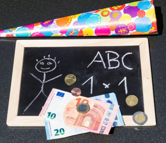 Eine kleine Tafel mit einem Strichmännchen und Schulaufgaben aus der ersten Klasse liegt neben einer Schultüte. Darunter liegen 30 Euro in Scheinen und etwas Kleingeld.