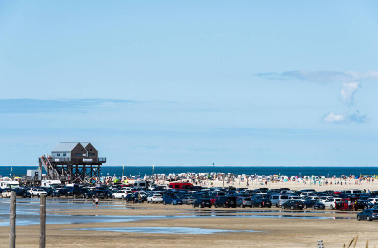 Ein weiter flacher Sand-Strand, auf dem viele Autos parken. Dahinter liegt das Meer. Ein kleines Holzhaus bietet Toiletten an. Urlauber sitzen in Strandkörben, gehen baden oder spielen Beach-Volleyball.