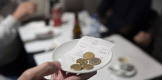 Eine Person hält ein kleines Schälchen in der Hand. In der Schale liegen Euro-Münzen. Im Hintergrund ist ein Tisch in einem Restaurant zu sehen.
