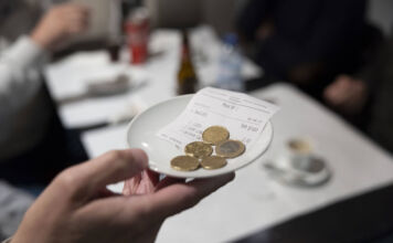 Eine Person hält ein kleines Schälchen in der Hand. In der Schale liegen Euro-Münzen. Im Hintergrund ist ein Tisch in einem Restaurant zu sehen.