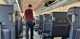 Ein Zugbegleiter der Deutschen Bahn trägt seine Arbeitskleidung und geht durch die Reihen eines Zugabteils. Ein Rucksack befindet sich auf der Ablage über den Sitzen.