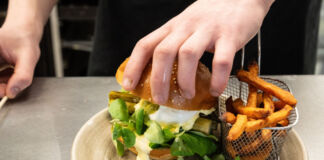 Die Hände eines Kochs sind gerade dabei, einen Burger mit Spargel zuzubereiten. Dieser liegt auf einem Teller. Daneben befindet sich ein Gitterkorb mit Pommes.