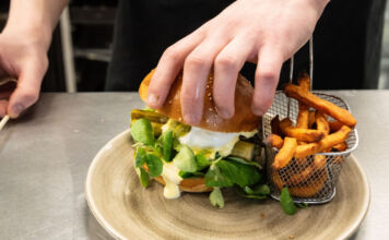 Die Hände eines Kochs sind gerade dabei, einen Burger mit Spargel zuzubereiten. Dieser liegt auf einem Teller. Daneben befindet sich ein Gitterkorb mit Pommes.