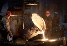 In einer Gießerei schütten zwei Arbeiter aus einem riesigen Kessel heißen flüssigen Stahl in einen anderen Behälter. Zum Schutz vor der Hitze tragen sie spezielle Handschuhe.