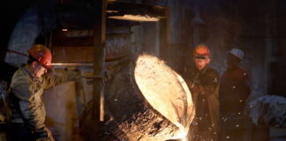 In einer Gießerei schütten zwei Arbeiter aus einem riesigen Kessel heißen flüssigen Stahl in einen anderen Behälter. Zum Schutz vor der Hitze tragen sie spezielle Handschuhe.