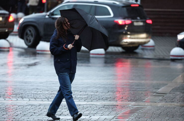 Eine Frau geht an einem regnerischen Tag durch die Stadt. In ihren Händen hält sie einen Regenschirm gegen Regen und Wind. Sie möchte sich vor der Nässe schützen. Im Hintergrund fahren Autos.