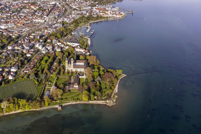 Ein Zeppelin fliegt über den Bodensee. Eine Luftaufnahme, auf der man den großen See und das Ufer sieht. Guter Blick auf Bäume, Häuser und den See.