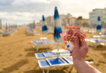 Die Hand eines Mannes hält mehrere 10-Euro-Scheine in die Kamera. Im Hintergrund verschwommen befinden sich an einem langen Strand zahlreiche Liegen mit zusammengeklappten Sonnenschirmen sowie zahlreiche Hotels.