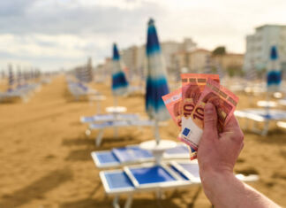 Die Hand eines Mannes hält mehrere 10-Euro-Scheine in die Kamera. Im Hintergrund verschwommen befinden sich an einem langen Strand zahlreiche Liegen mit zusammengeklappten Sonnenschirmen sowie zahlreiche Hotels.