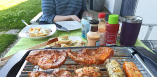 Auf einem Tisch auf dem Balkon steht ein Tischgrill, auf dem sich Fleisch und Grillkäse befinden. Dahinter stehen Ketchup und Grillsaucen. Eine Frau sitzt am Tisch und hat vor sich zwei Teller mit Essen und eine Getränke-Dose stehen.