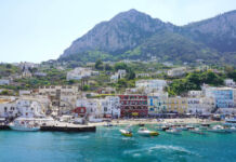 Capri - eine der beliebtesten Urlaubsinseln der Deutschen liegt direkt am Meer, mitten im sommerlichen Sonnenschein. Im Hintergrund liegen hohe Berge und grüne Wälder.