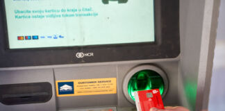 Eine rote Bankkarte wird in den Schlitz beim Automaten gesteckt. Der Bildschirm gibt Anweisungen an den Kunden, wie er den Automaten nutzen kann.
