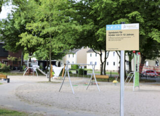 Ein leerer, sauberer Spielplatz mit Schaukel und mehreren Gerüsten zum Spielen steht zwischen mehreren grünen Bäumen und einer Häuserzeile im Hintergrund friedlich an einem sonnigen Tag und wartet auf spielende Kinder, während ein Schild auf die Regeln hinweist.