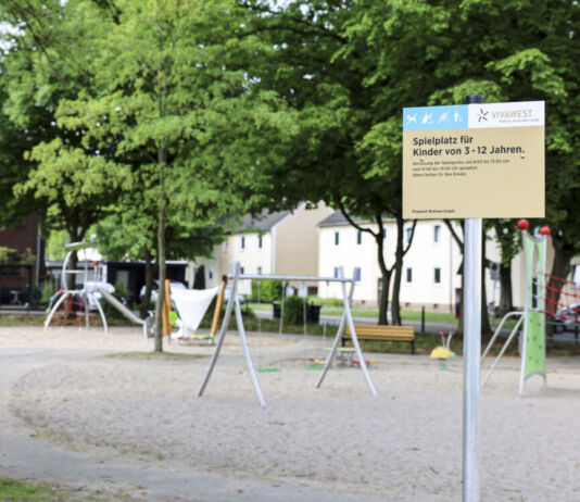 Ein leerer, sauberer Spielplatz mit Schaukel und mehreren Gerüsten zum Spielen steht zwischen mehreren grünen Bäumen und einer Häuserzeile im Hintergrund friedlich an einem sonnigen Tag und wartet auf spielende Kinder, während ein Schild auf die Regeln hinweist.