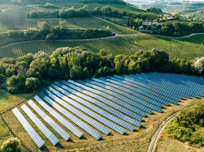 Auf einer Luftaufnahme eines Solarparks auf dem Land sind zahlreiche Solarpanels zu sehen, die umringt von Bäumen mitten in der Landschaft auf einer grünen Wiese in der Nähe eines Dorfes stehen und Strom und Sonnenenergie liefern. Eine Solarfirma rutscht nun in die Insolvenz.