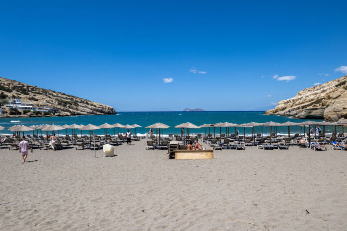 An einem Strand stehen viele Liegen mit Schirmen aus Stroh. Dahinter ist das türkise Meer zu sehen. Der Himmel ist blau und die Sonne scheint.