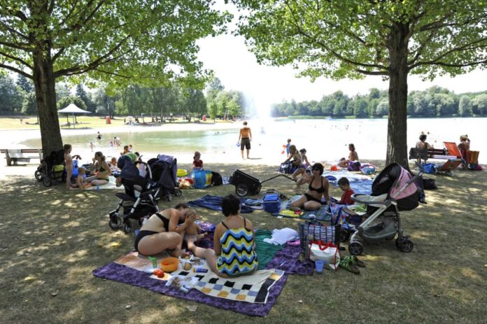 Zahlreiche Badegäste liegen auf vielen bunten Decken an einem Badesee bei schönem, sonnigen Wetter. Sie liegen im Schatten, andere Gäste gehen in das Wasser der warmen Seen baden.