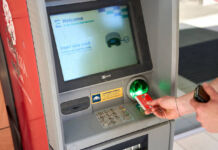 Eine Hand steckt eine Girokarte in einen Geldautomaten der Sparkasse, um Geld abzuheben. Auf dem Bildschirm steht in englischer Sprache, dass man seine Karte einstecken soll.